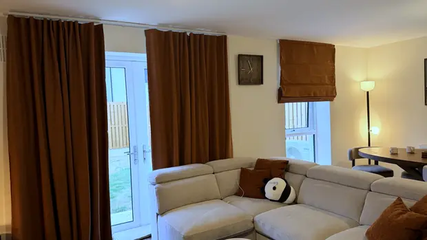 Living room with rust-coloured full-length curtains and a matching Roman blind above a corner sofa.